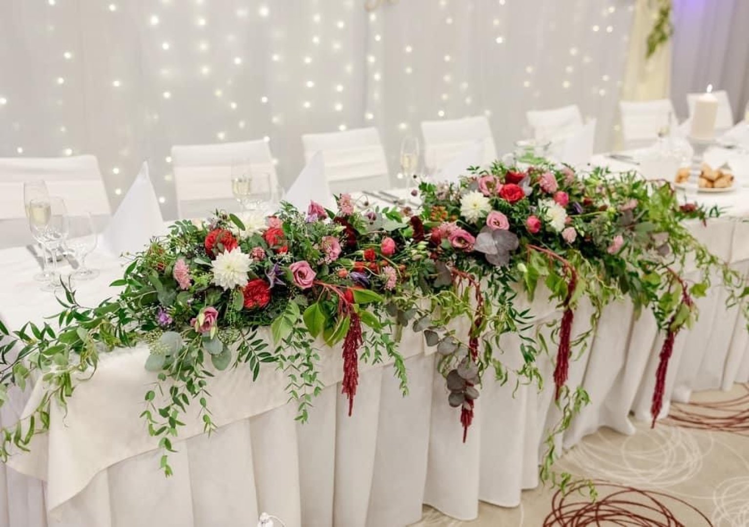 Elegantná kvetinová výzdoba na hlavnom svadobnom stole v rámci aranžérskych a floristických služieb s bielym pozadím a svetielkami.