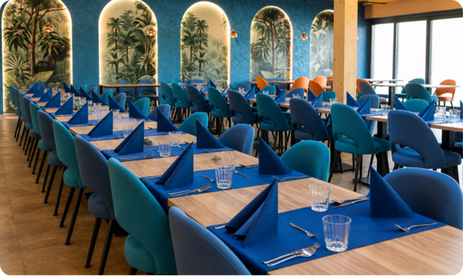 Interiér reštaurácie Restaurant AZURO s prestretými stolmi s modrými obrusmi a dizajnovými tyrkysovými stoličkami pred stenou s tropickými motívmi.