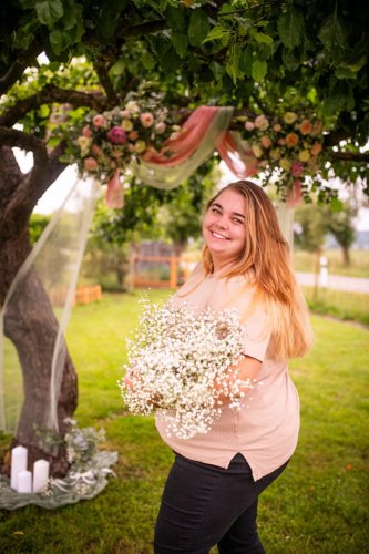 Usmievavá floristka z HAHADecor drží veľkú kyticu bielych kvetov pred vyzdobeným svadobným stromom v záhrade