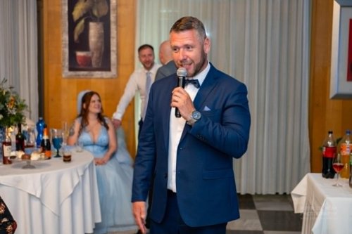 Svadobný moderátor Martin Matuška v modrom obleku s mikrofónom v ruke počas príhovoru na svadobnej hostine.