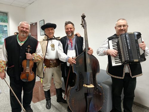 Traja usmiati členovia ľudovej hudby Halúzkovci v tradičných krojoch pózujúci za dreveným rámom.