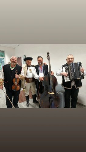 Traja usmiati členovia ľudovej hudby Halúzkovci v tradičných krojoch pózujúci za dreveným rámom.