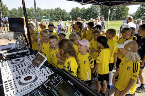 Skupina detí v žltých dresoch so záujmom sleduje DJ techniku počas kultúrnej akcie v exteriéri.