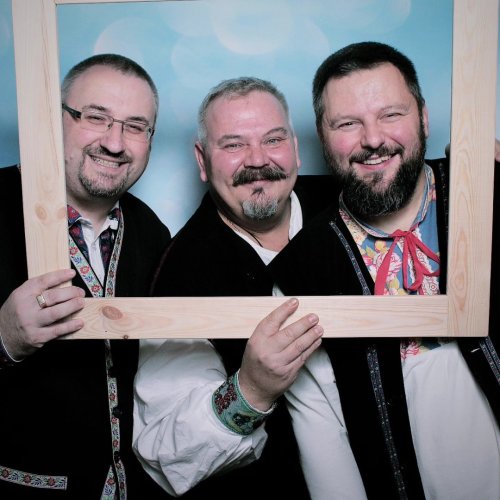 Traja usmiati členovia ľudovej hudby Halúzkovci v tradičných krojoch pózujúci za dreveným rámom.