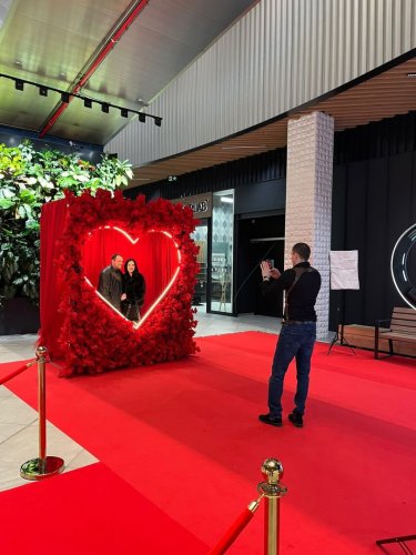 Pár pózuje v Love Story kútiku pred svietiacim srdcom z červených kvetov na červenom koberci, zatiaľ čo ich fotograf fotí.