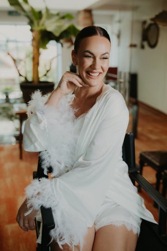 Usmievajúca sa nevesta v elegantnom bielom župane s páperím počas ranných príprav, zachytená fotografom Adriánom Juríčkom.