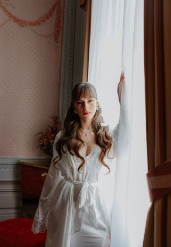 Elegantná nevesta v hodvábnom župane pripravujúca sa na svadbu pri okne v historickom interiéri.