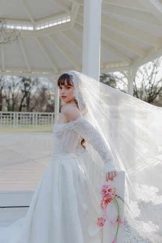 Elegantná nevesta v čipkovaných svadobných šatách s dlhým závojom pózuje v bielom altánku počas svadby.