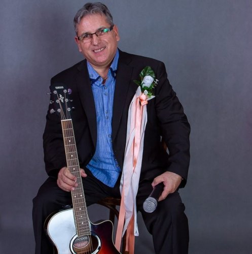 Usmievavý starejší s gitarou v obleku s bielou svadobnou pierkom, držiaci mikrofón.