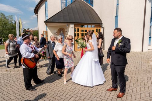 Starejší s gitarou v ľudovom klobúku hrá novomanželom pred kostolom počas gratulácií hostí.