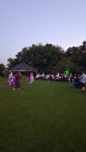 Hostia sediaci pri stoloch na trávniku počas večernej svadobnej oslavy vonku so stanom a stromami v pozadí.